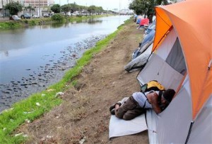 In this April 22, 2015 photo, Derek Villanueva, who is homeless, sleeps outside a friends tent in an encampment along a canal in Honolulu. The Honolulu Star-Advertiser reported that the city council voted Wednesday, May 6, to expand measures restricting people from sitting and lying down despite questions about their legality. (AP Photo/Cathy Bussewitz)